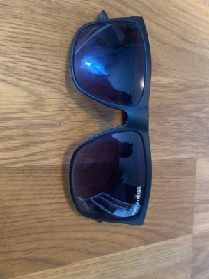 Svarta solglasögon från Emporio Armani - Stilrena svarta solglasögon från Emporio Armani med bred båge och rektangulära glas. Snygg logga på skalmen och mörka glas som ger en cool look. Perfekta för att lyfta din outfit under soliga dagar.
