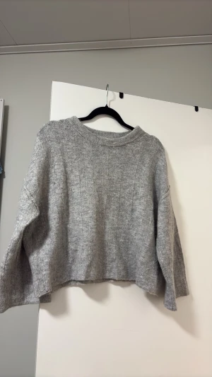 Grå oversize stickad tröja - Mysig grå stickad tröja i oversize-modell med breda ärmar och rund hals. Tröjan har en diskret ribbad struktur och är tillverkad i ett mjukt material som passar perfekt för kyliga dagar.