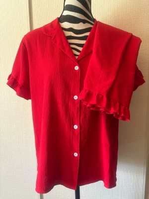 Röd pyjamas med volangdetaljer - Säljer ett set med röd pyjamas i mjukt tyg. Skjortan har korta ärmar, vita knappar och volang vid ärmslut. Byxorna har resår i midjan och volang längst ner vid bensluten. Perfekt för mysiga kvällar hemma.
