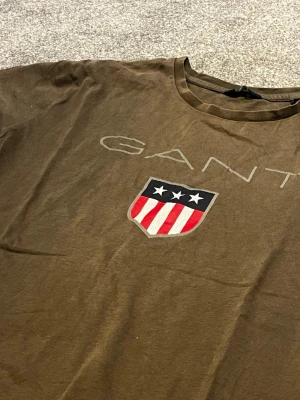 Oliv Grön GANT t-shirt med logga - Oliv grön t-shirt från GANT med rund hals och korta ärmar. På bröstet finns ett stort GANT-tryck och en sköld med stjärnor och rödvita ränder. Materialet ser ut att vara bomull och modellen är klassisk och rak. Denna t-shirt passar personer som är ca 160 cm till 180 cm lång. vid minsta intresse eller fundering var vänligen att meddela frågor. Med vänliga hälsningar Eric 