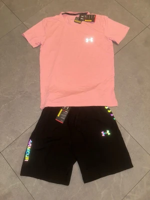  Rosa t-shirt & svarta shorts - Säljer ett sportigt set med en ljusrosa t-shirt och svarta shorts. T-shirten har rund hals och logga på bröstet, shortsen har resår i midjan och färgglad logga samt texten 'ARMOUR' på benet. Perfekt för träning eller chill.