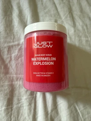 Just Glow Watermelon Explosion bodyskrubb - Oöppnad och plomberad. Sockerbaserad bodyskrubb från Just Glow med doft av vattenmelon. Kommer i en stor burk med vit skruvlock och har en härligt rosa färg. Innehåller sheasmör och vitamin E för extra återfuktning. Tillverkad i Sverige. Nypris 399kr