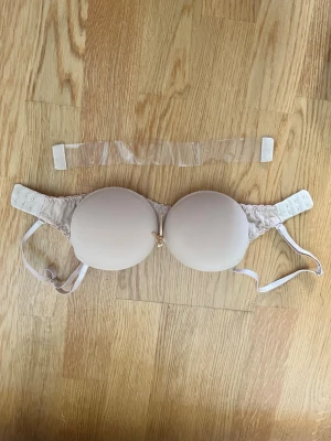 Beige push up  axelbandslös bh med silikonband vid ryggen - Säljer en beige axelbandslös bh med avtagbara axelband och  silikonband vid ryggen för bättre stöd. Bh:n har vadderade kupor och en liten rosett framtill. Materialet är mjukt och slätt, perfekt under ljusa plagg. Oanvänd endast tvättad