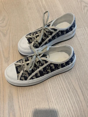Christian Dior Oblique sneakers låga - Snygga låga sneakers från Christian Dior med klassiskt blått och vitt Oblique-mönster. Skorna har vita snören, vit gummisula och detaljer med logotyp på plösen. Tillverkade i canvas med robust sula för en trendig look. Skorna är köpta secondhand. Äkthets bevis eller kvitto finns ej. (Skorna är 39 men skulle säga att det passar bättre på en 38) 