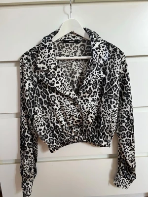 Leopardmönstrad croppad skjorta - Snygg under stängd kavaj/blazer. Säljer en croppad skjorta med svart och vit leopardmönster. Skjortan har långa ärmar, bred krage och resår i midjan för en snygg passform. 