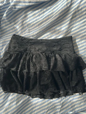 Svart volangkjol i spets  - Supersöt svart spets kortkjol med dubbla volanger. Kjolen har en bred midja och även inbyggada shorts så man slipper oroa sig över att kjolen flyger upp! Perfekt nu inför sommaren! Helt oanvänd!🥰storlek xs men skulle säga att den är mer än som en xxs!