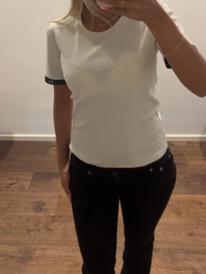 Vit t-shirt med svarta detaljer - Vit t-shirt med rund hals och svarta detaljer med små nitar på ärmsluten. T-shirten har en klassisk passform och är perfekt för en clean och stilren look. Materialet känns mjukt och bekvämt, troligtvis bomull.