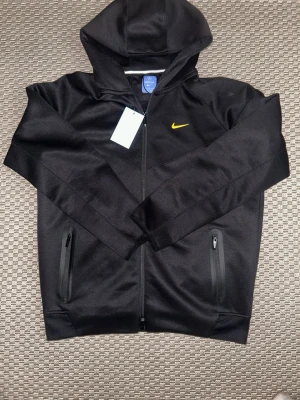 Svart Nike hoodie - Snygg svart hoodie från Nike med dragkedja och gul swoosh-logga på bröstet. Hoodien har två fickor med dragkedja och en klassisk huva. Tillverkad i ett mjukt och sportigt material som passar perfekt till streetwear-stilen.