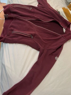 Vinröd hoodie med dragkedjor - Säljer en vinröd hoodie med flera dragkedjor framtill och på fickorna. Hoodien har en patch på ärmen och är tillverkad i mjukt material, perfekt för chill dagar. Snygg och enkel design som passar till det mesta.