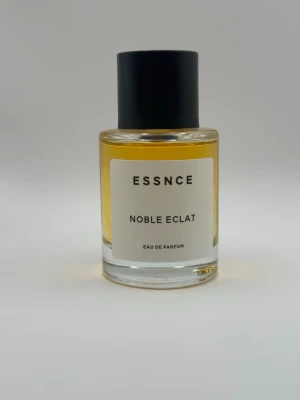 ESSNCE Noble Eclat Eau de Parfum - ESSNCE Noble Eclat Eau de Parfum i en stilren, rund glasflaska med svart lock. Parfymen har en gyllengul vätska och etiketten är vit med svart text. Flaskan ger ett modernt och minimalistiskt intryck, perfekt för dig som gillar enkel och elegant design.