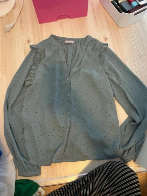 Blå prickig blus från Aware - Söt ljusblå blus från Aware med svarta små prickar och volangdetaljer vid axlarna. Blusen har långa ärmar och knäppning framtill. Tunt och luftigt material som ger en avslappnad känsla.