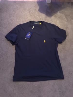 Marinblå t-shirt från Polo Ralph Lauren - Snygg marinblå t-shirt från Polo Ralph Lauren med klassisk gul broderad logga på bröstet. T-shirten har rund halsringning och korta ärmar. Tillverkad i mjuk bomull som känns skön mot huden. Perfekt till jeans eller shorts för en clean look.