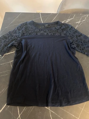 Mörkblå topp med spetsdetaljer - Snygg mörkblå topp från Lindex med spets över axlar och ärmar. Toppen har rund halsringning och halvtransparent spetsmönster upptill samt på ärmarna. Perfekt för dig som gillar feminina detaljer och enkel stil.