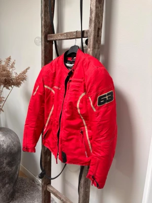 Röd MC-jacka från Bikers - Korallröd motorcykeljacka från Bikers med svarta och beige detaljer. Jackan har dragkedja framtill, två fickor med dragkedja. Tillverkad i slitstarkt textilmaterial och har sportig design med markerade sömmar. Inbyggda skydd för axlar & armbågar. Innerjackan kan man ta loss enkelt med dragkedja 