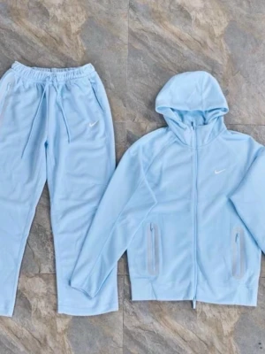 Nike NOCTA ljusblå set - Nike NOCTA tracksuit set i ljusblått med hoodie och matchande byxor. Hoodien har dragkedja, fickor och NOCTA-detaljer, medan byxorna har dragsko i midjan och en liten Nike-logga. Tillverkat i mjukt och bekvämt material.