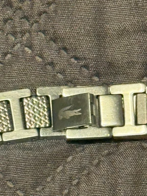 Lacoste armband - Säljer ett stilrent armband i silverfärg från Lacoste med klassisk krokodil-logga på låset. Armbandet har breda länkar med både släta och mönstrade ytor som ger en cool och modern look. Perfekt för dig som gillar att sticka ut med accessoarer.