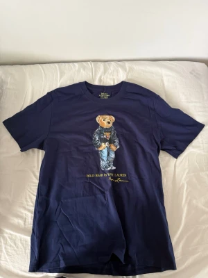 Marinblå Polo Bear t-shirt Ralph Lauren - Snygg marinblå t-shirt polo Bear  Ralph Lauren med ikoniska Polo Bear-trycket på bröstet. T-shirten har rund hals och korta ärmar. Tillverkad i mjuk bomull för skön känsla och avslappnad passform. Perfekt för dig som gillar klassisk streetstyle.