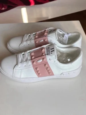 Vita sneakers med rosa nitar från Valentino - Snygga vita sneakers från Valentino med bred rosa rem över vristen dekorerad med pyramidformade nitar. Skorna har en klassisk låg siluett, vita snören och detaljerade sömmar. Tillverkade i slätt läder för en clean och modern look.