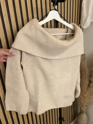 Beige stickad offshoulder tröja - Mysig beige stickad tröja med bred, nedvikt krage och långa ärmar. Tröjan har en avslappnad passform och är ribbstickad i ett mjukt material. Perfekt för kyliga dagar och enkel att styla till olika outfits.