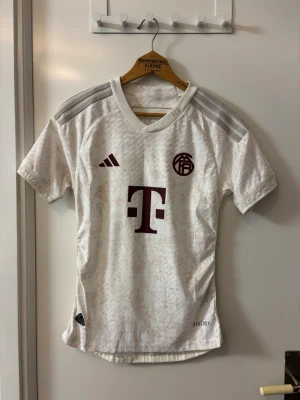 Vit Adidas fotbollströja FC Bayern - Vit fotbollströja från Adidas med FC Bayern München-logga och Telekom-sponsor i vinrött. Tröjan har diskret blommönster, gråa axelränder och HEAT.RDY-teknologi för extra ventilation. Perfekt för träning eller match.