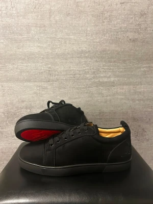 Svarta Louboutin sneakers med röd sula - Snygga svarta sneakers från Christian Louboutin med klassisk röd sula. Skorna har lågt skaft, svarta skosnören och guldfärgad insida. Tillverkade i mocka och läder för en lyxig känsla. Perfekta för dig som vill sticka ut med exklusiva detaljer.