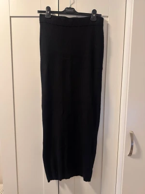Svart stickad långkjol - En svart, stickad långkjol med rak passform och bred resår i midjan. Kjolen är enkel och stilren, perfekt för dig som gillar en clean look. Materialet är mjukt och stretchigt vilket gör den bekväm att bära hela dagen.