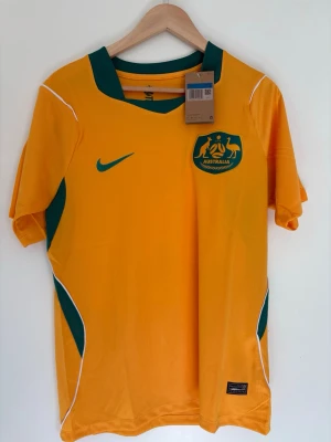 Australien Nike matchtröja gul M - Säljer en officiell gul matchtröja från Nike för Australiens landslag. Tröjan har gröna detaljer, landslagets emblem på bröstet och Dri-FIT-material som andas. Perfekt för fotboll eller träning. Klassisk V-ringning och korta ärmar.