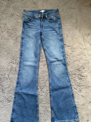 Blå bootcut jeans med broderade fickor - Ett par klassiska blå bootcut jeans med lätt tvättad look. Jeansen har fem fickor och snygga broderade detaljer på bakfickorna. Tillverkade i denim med normal midja och dragkedja framtill. Perfekta för en avslappnad och trendig stil.