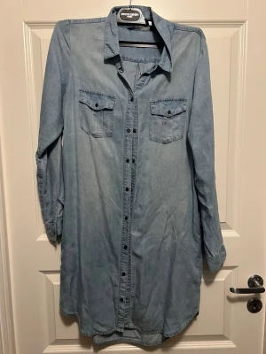 Ljusblå jeansklänning från Vero Moda - Ljusblå jeansklänning med lång ärm och knappar hela vägen framtill. Två bröstfickor med lock och klassisk skjortkrage. Perfekt för en avslappnad och trendig look. Har ett nästan osynligt litet märke på ryggen.