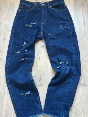 Vintage archive Mörkblå distressed jeans från ONLY - Mörkblå jeans med relaxed fit och flera slitningar framtill och baktill. Klassisk femficksmodell med hög midja och råa benslut. Jeansen är tillverkade i kraftig denim och har en trendig, avslappnad look.