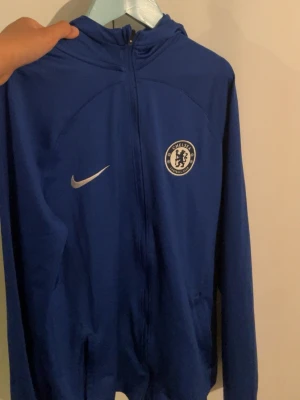 Chelsea FC blå Nike träningsjacka - Chelsea FC träningsjacka i blått med dragkedja framtill och huva. Jackan har Chelsea Football Club-emblem på bröstet och Nike-logga i vitt. Tillverkad i ett lätt och sportigt material, perfekt för träning eller matchdag.