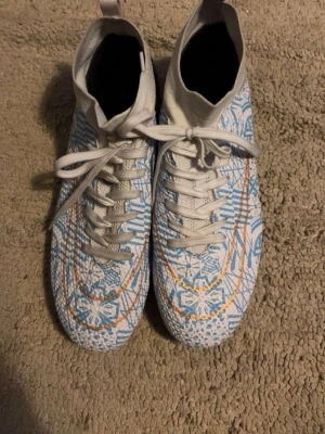 Nike vita och blå fotbollsskor - DOM TVÄTTAS INNAN! Säljer ett par Nike fotbollsskor med högt skaft i vitt med blått och orange grafiskt mönster. Skorna har snörning och dobbar i blått och vitt för bra grepp på planen. Perfekta för dig som vill sticka ut på matchen!