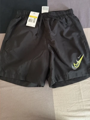 Svarta badbyxor från Nike - Snygga svarta badbyxor från Nike med elastisk midja och dragsko. På vänster ben finns en stor gul och vit Nike-logga. Badbyxorna har en bakficka och är tillverkade i snabbtorkande material, perfekta för stranden eller poolen.