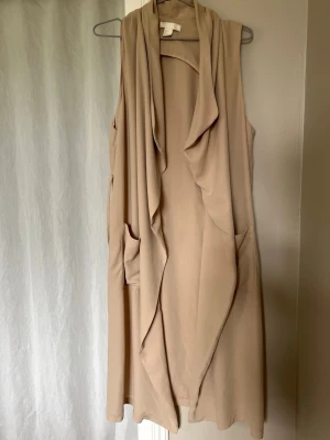 Beige lång kavajväst med fickor - Stilren beige kavajväst med draperad framsida och två fickor. Västen är lång och har en öppen design utan knappar eller dragkedja. Perfekt att slänga över en t-shirt eller skjorta för en avslappnad men snygg look.