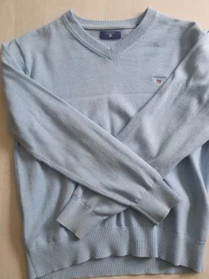 Ljusblå stickad tröja från Gant - Snygg ljusblå stickad tröja från Gant med V-ringning och diskret logga broderad på bröstet. Tröjan har långa ärmar, ribbade muddar och är tillverkad i mjukt material som känns skönt mot huden. Perfekt för dig som gillar klassisk och stilren look.