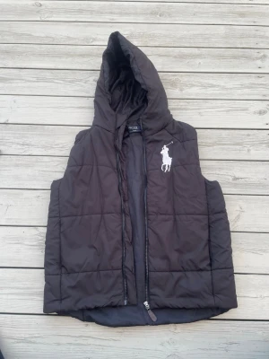 Svart dunväst från Polo Ralph Lauren - Snygg svart dunväst från Polo Ralph Lauren med stor vit broderad logga på bröstet. Västen har huva och dragkedja framtill. Perfekt för lager på lager och ger en sportig vibe. Tillverkad i vadderat material som håller värmen.