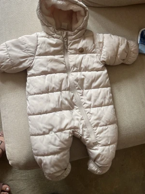dunoverall med huva - Säljer en beige dunoverall för baby med mysig huva och mjukt foder. Overallen har dragkedja framtill som går snett över kroppen och täckta fötter. Perfekt för kalla vinterdagar och håller värmen riktigt bra.