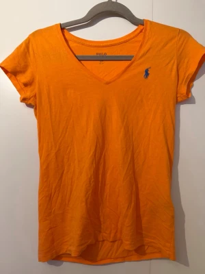 Orange v-ringad t-shirt från Polo Ralph Lauren - Säljer en orange t-shirt från Polo Ralph Lauren med v-ringning och korta ärmar. T-shirten har en blå broderad logga på bröstet och är gjord i mjuk bomull. Perfekt för dig som gillar klassiska och färgstarka plagg. OBS den har defekter på ena sidan armen 😊