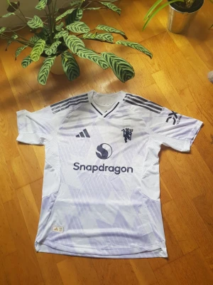 Vit Manchester United fotbollströja (strl L) - Snygg vit Manchester United fotbollströja från Adidas med lila mönster, mörklila detaljer och Snapdragon-logga på bröstet. Tröjan har V-ringning, tre ränder på axlarna och klubbmärke på vänster sida. Tillverkad i lätt och ventilerande material.