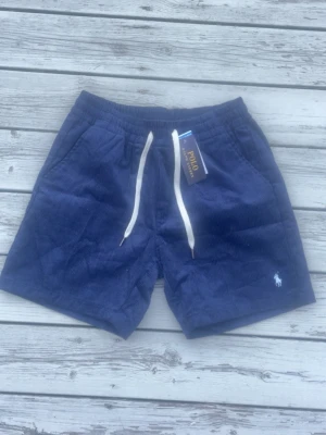 Blå manchester shorts från Polo Ralph Lauren - Säljer ett par mörkblå manchester shorts från Polo Ralph Lauren med vit snörning i midjan och klassisk logga broderad på benet. Shortsen har en relaxed passform och är perfekta för sommardagar. Snyggt ribbade detaljer och två sidofickor.