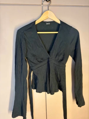 Svart omlottopp från Gina Tricot - Snygg svart omlottopp från Gina Tricot med långa ärmar och knytband i midjan. Toppen har en djup v-ringning och är tillverkad i ett lätt, strukturerat tyg som ger en cool look. Perfekt att styla med jeans eller kjol.