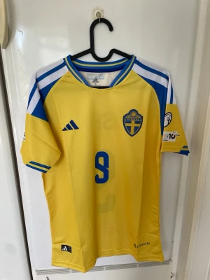 Sverige matchtröja Adidas gul/blå - Säljer en officiell Sverige matchtröja från Adidas i gult med blå detaljer och nummer 9 på bröstet. Tröjan har korta ärmar, blå och vita ränder på axlarna samt landslagets emblem på vänster bröst. Tillverkad i lätt och ventilerande material, perfekt för fotboll eller träning.