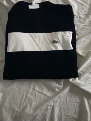 Svart tröja med vit rand från Lacoste - Snygg svart tröja från Lacoste med bred vit rand över bröstet och längs ärmarna. Klassisk rund halsringning och den ikoniska gröna krokodilloggan på bröstet. Perfekt för dig som gillar stilrena och sportiga plagg.