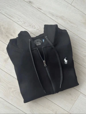 Svart hoodie från Polo Ralph Lauren - Svart hoodie med dragkedja från Polo Ralph Lauren. Storlek S, tags med följer!,  Klassisk design med vit broderad logga på bröstet, huva med dragsko och ribbade muddar. Tillverkad i mjukt bomullsmaterial som är skönt att ha på sig. Perfekt för en avslappnad och stilren look. 