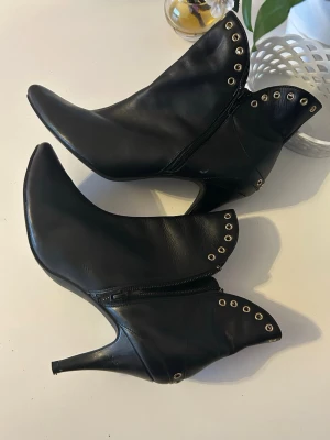 Svarta stövletter med nitar och spetsig tå - Snygga svarta stövletter i skinnimitation med spetsig tå och smal klack. Dekorativa guldfärgade nitar längs kanten ger en edgy look. Dragkedja på insidan för enkel på- och avtagning. Perfekta för dig som vill sticka ut lite extra. stövletter i skinnimitation