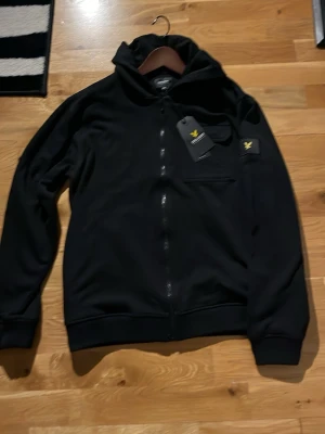 Svart hoodie från Lyle & Scott - Snygg svart hoodie från Lyle & Scott med dragkedja framtill och klassisk huva. Har en liten gul logga på ärmen och bröstet samt ribbade muddar. Perfekt för en chill och stilren look.