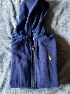 Hoodie från Polo Ralph Lauren - Säljer en marinblå hoodie från Polo Ralph Lauren med dragkedja och huva. Klassisk gul broderad logga på bröstet. Perfekt för en avslappnad och stilren look! Dock hål på begge armbågar, se bild!!!