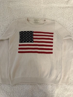 Beige stickad tröja med USA-flagga - Stickad beige tröja från Playback med stor amerikansk flagga i rött, vitt och blått på bröstet. Tröjan har rund halsringning och ribbade muddar vid ärmslut och nederkant. Perfekt för dig som gillar statement-plagg och vill sticka ut.