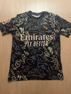 Real Madrid Benzema matchtröja - Säljer en svart Real Madrid matchtröja från Adidas med graffitiinspirerat mönster i vitt och blått. Tröjan har klubbmärke, sponsortryck och nummer 9 med Benzema på ryggen. Tillverkad i lätt och ventilerande material, perfekt för fotbollsträning.
