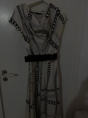 Vit långklänning med kedjemönster - Vit långklänning med svart kedjemönster och bred svart bälte med guldfärgat spänne. Klänningen har korta ärmar och en draperad halsringning. Perfekt för dig som vill sticka ut med en unik och trendig look.